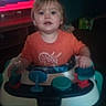 toddler, child, baby, sitting, activity_walker, toy, orange_shirt, indoors, living_room, television, portrait, face, eyes, looking_at_camera, curious, hands, colorful, dim_lighting, blurry, home