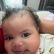 Delta is registered to the contest to win money with this photo: baby, infant, face, eyes, tongue, curly_hair, clothing, white_outfit, patterned_clothing, indoor, closeup, smiling, cute, person, child, skin, head, expression, comfort, lying_down