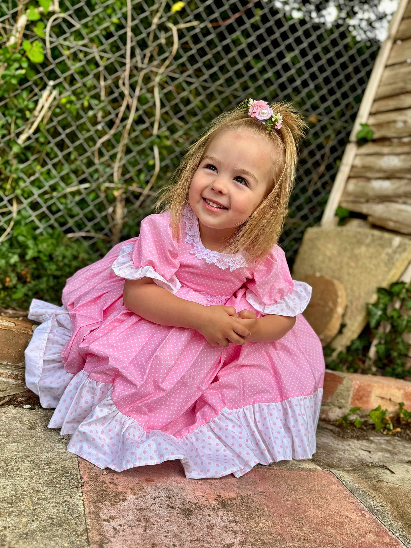 Dottie joined the competition — help win amazing prizes! child, girl, smiling, dress, pink, polka_dot, hair_accessory, flower, sitting, outdoor, garden, fence, greenery, concrete, stone, happy, portrait, cute, person, young