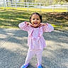 child, toddler, pink_clothing, outdoor, smiling, playful_pose, paved_path, white_fence, greenery, sunny_day, happy, girl, casual_shoes, braided_hair, cute, daylight, portrait, fashion, young_child, nature