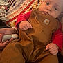 baby, child, overalls, red_shirt, crocheted_blanket, hand, face, expression, furniture, indoor, person, toy, clown_doll, plush, couch, casual_clothing, portrait, infant, closeup, human