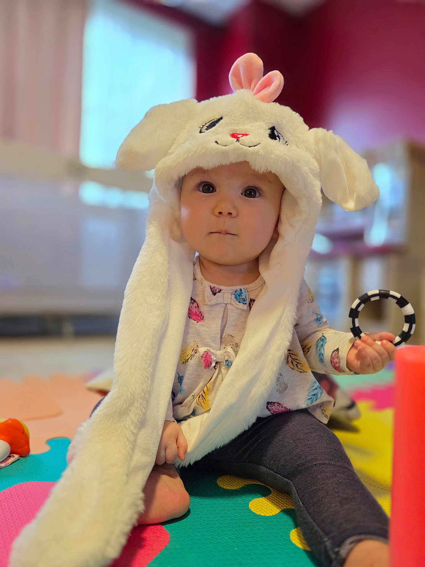 Noelle is registered to the contest to win money with this photo: baby, child, indoor, play_mat, toy, bunny_hat, cute, fluffy, costume, sitting, colorful, infant, face, expression, legging, foot, hand, clothing, room, soft_focus