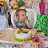 toddler, child, birthday_cake, high_chair, messy_face, green_dress, balloons, giraffe_decoration, gift_bags, party, celebration, cake, table, face, hands, colorful, decorations, indoor, smudged_icing, happy