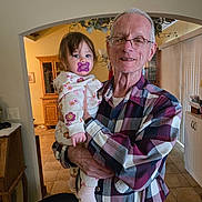 Brayleanna joined the competition — help win amazing prizes! elderly_man, toddler, pacifier, plaid_shirt, glasses, indoor, home, cabinet, plants, tile_floor, holding, child, portrait, family, smiling, person, clothing, socks, light, door