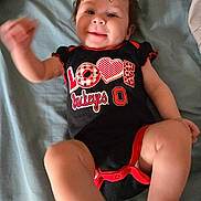 Brayleanna joined the competition — help win amazing prizes! arm, baby, bed, blanket, casual, child, clothing, cute, face, happy, indoors, infant, leg, lying_down, onesie, person, red_black, smiling, text_on_clothing, waving