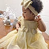 toddler, child, dress, yellow_dress, headband, curly_hair, table, wooden_table, indoor, decor, pineapple_decor, ornamental_feathers, sitting, barefoot, girl, portrait, cute, smiling, playful, accessory
