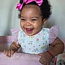 baby, girl, smiling, pink_bow, curly_hair, crib, blanket, tulle_sleeves, happy, indoor, portrait, child, cute, infant, patterned_clothing, red_bracelet, furniture, home, face, playpen