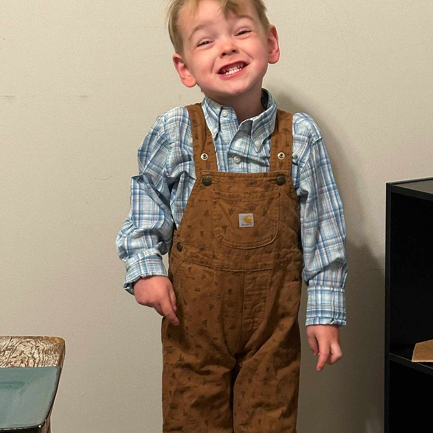 Waylon joined the competition — help win amazing prizes! boy, casual_clothing, child, expression, furniture, happy, home, indoor, light_skin, missing_teeth, overalls, plaid_shirt, portrait, real_person, shelf, short_hair, smile, standing, wall, wooden_table