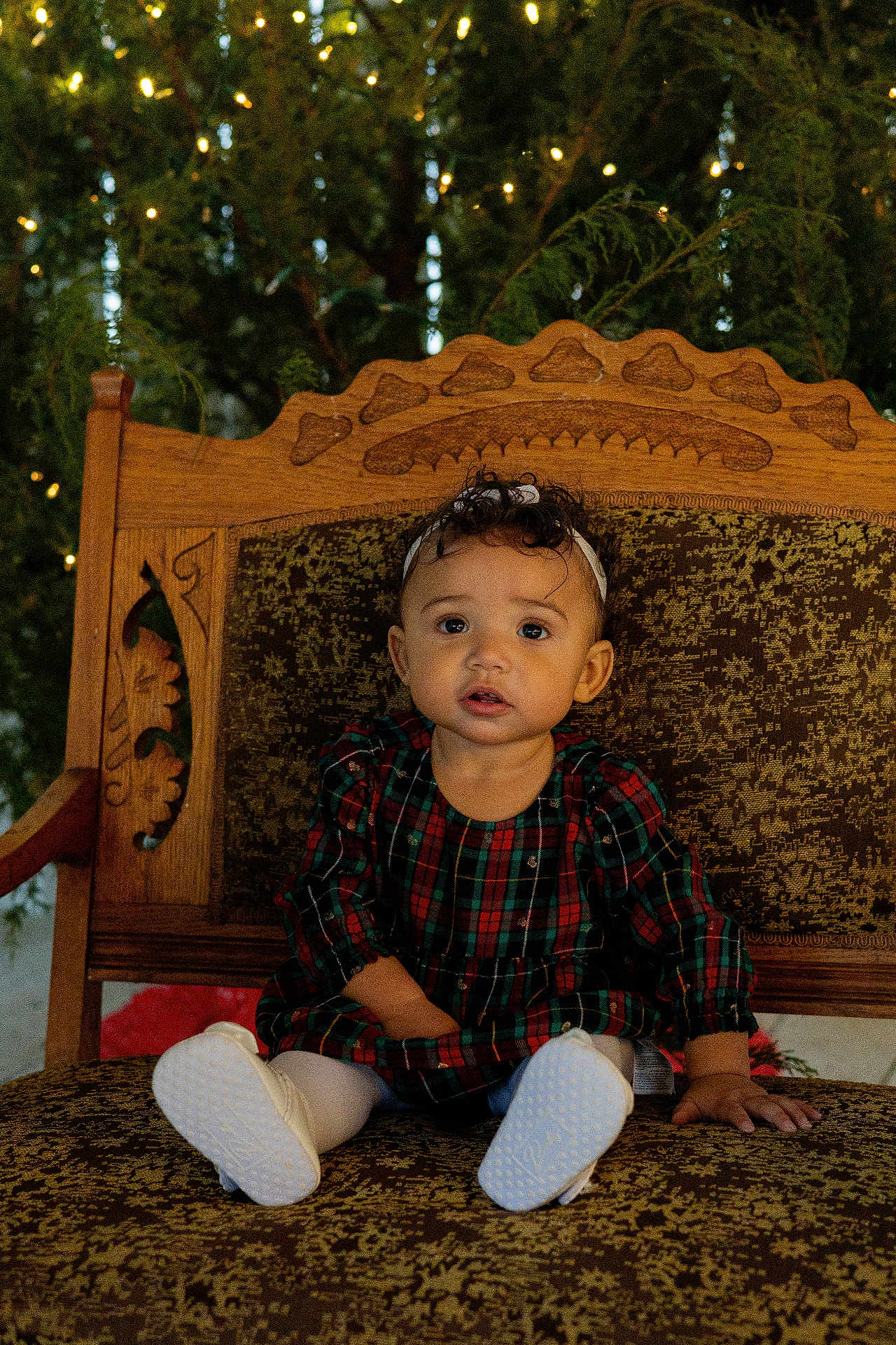 Ryia joined the competition — help win amazing prizes! toddler, child, plaid_dress, white_shoes, headband, chair, wooden_chair, patterned_upholstery, curly_hair, indoors, holiday_lights, greenery, portrait, sitting, cute, festive, expression, young_child, baby, holiday