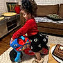 Razen participe au concours pour gagner de l'argent avec cette photo : black_skirt, brown_couch, cake, carpet, child, curly_hair, decorative_pillow, heart_pattern, indoor, lion_slippers, living_room, plant, playful, red_shirt, slippers, table, toddler, toy_bike, wall_art, wooden_floor