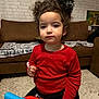 bow, brick_wall, carpet, casual_clothing, child, couch, curly_hair, curly_hair_bow, expressionless, furniture, home, indoor, living_room, person, playtime, portrait, red_shirt, toddler, toy_vehicle, young_child