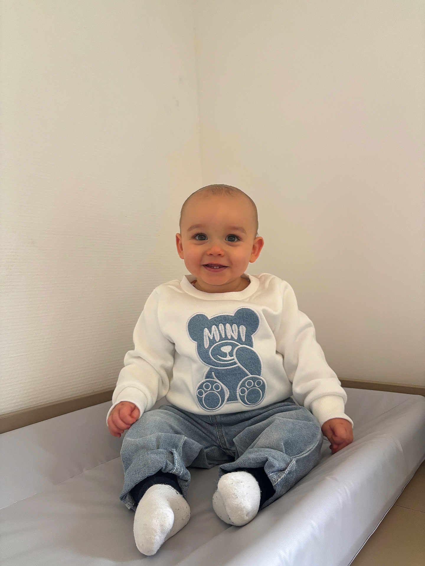 Kaïs Nomé participe au concours pour gagner de l'argent avec cette photo : baby, child, smiling, sitting, indoor, clothing, sweatshirt, jeans, socks, face, head, person, cute, happy, teddy_bear, padded_surface, wall, minimalist, portrait, casual