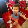 toddler, child, red_chair, blue_top, orange_skirt, white_shoes, curly_hair, smiling, indoor, tile_floor, person, cute, sitting, baby, happy, face, looking_up, casual_clothing, restaurant, bright_colors