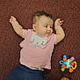 baby, child, pink_shirt, cat_design, carpet, toy, colorful_toy, navy_pants, curly_hair, earrings, lying_down, arms_up, feet, brown_shoes, floor, indoor, cute, infant, person, casual_clothing