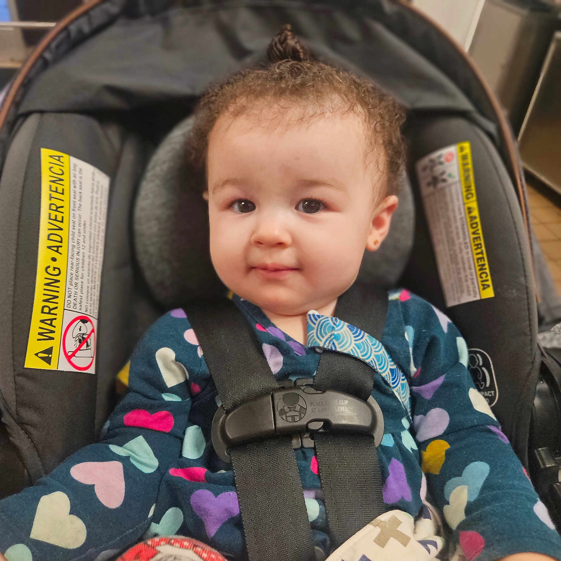 Amber is registered to the contest to win money with this photo: baby, car_seat, child, infant, safety_harness, heart_pattern, clothing, curly_hair, topknot, indoors, warning_label, seat_belt, cute, smile, person, portrait, young_child, face, straps, secure