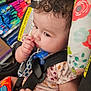 attention, baby, car_seat, child, closeup, clothing, curly_hair, cushion, cute, face, floral_pattern, hand, indoor, infant, pacifier, portrait, seatbelt, secure, sitting, warning_label