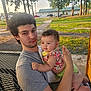 baby, bench, casual_clothing, child, curly_hair, daylight, family, grass, greenery, holding, infant, man, nature, outdoor, park, people, portrait, sidewalk, table, trees
