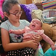 Savannah joined the competition — help win amazing prizes! baby, blanket, child, colorful, couch, cozy, face, family, glasses, holding, home, indoor, patterned_pants, person, pillow, pink_shirt, seated, sibling, smiling, toddler