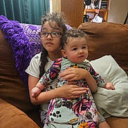 Savannah is registered to the contest to win money with this photo: child, baby, glasses, couch, purple_blanket, floral_dress, braid, indoor, sitting, family, cute, curly_hair, holding, cushion, brown_furniture, portrait, casual_clothing, home, two_children, smile