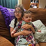 child, baby, glasses, couch, purple_blanket, floral_dress, braid, indoor, sitting, family, cute, curly_hair, holding, cushion, brown_furniture, portrait, casual_clothing, home, two_children, smile