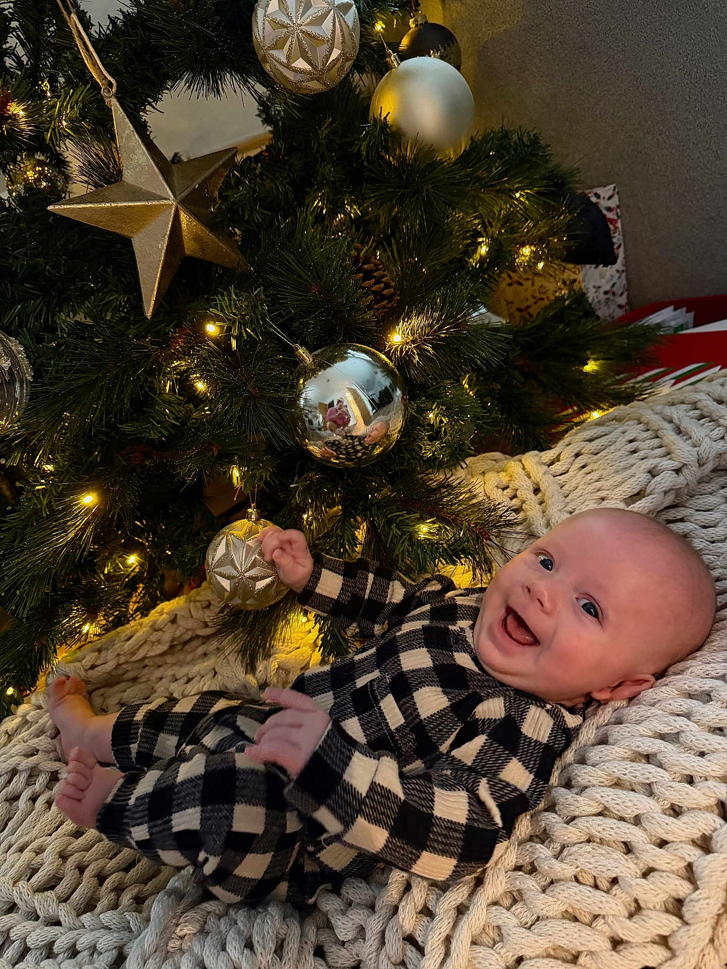 Sonny is registered to the contest to win money with this photo: baby, child, christmas_tree, ornaments, lights, blanket, knit, smiling, happy, holiday, festive, indoor, decorations, gold, silver, star, person, infant, cozy, plaid