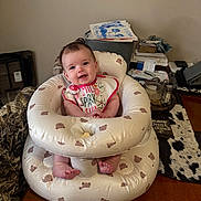 Blakley is registered to the contest to win money with this photo: baby, infant, smiling, inflatable_seat, bib, table, papers, household_items, clutter, indoor, person, face, feet, wooden_table, decor, happy, child, cute, seated, room