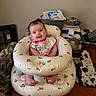 baby, infant, smiling, inflatable_seat, bib, table, papers, household_items, clutter, indoor, person, face, feet, wooden_table, decor, happy, child, cute, seated, room