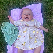 Peach is registered to the contest to win money with this photo: baby, smiling, dress, giraffe_pattern, blanket, grass, outdoor, happy, child, infant, person, cute, lying_down, playful, summer, nature, young, cheerful, clothing, pacifier_clip