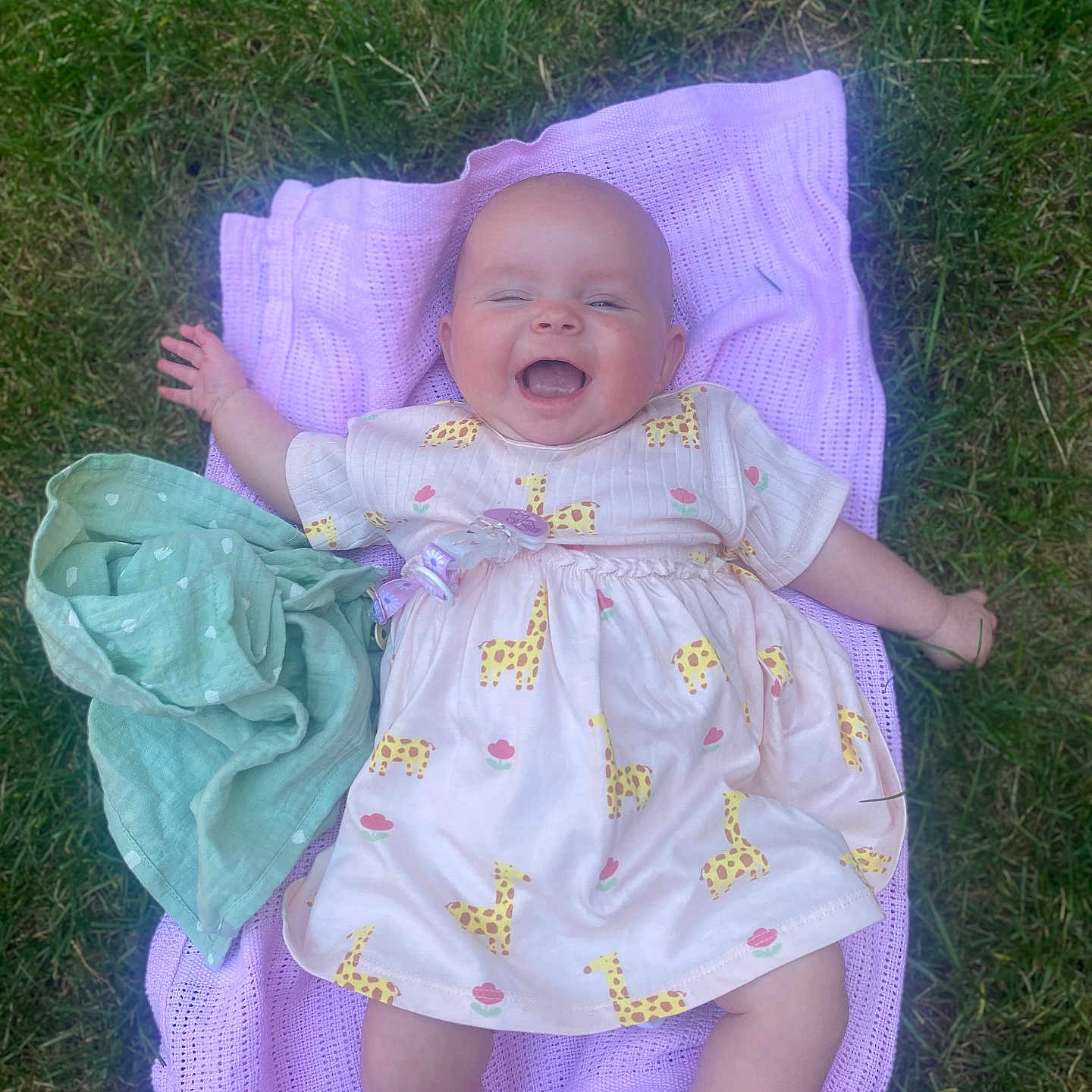 Peach is registered to the contest to win money with this photo: baby, blanket, cheerful, child, clothing, cute, dress, giraffe_pattern, grass, happy, infant, lying_down, nature, outdoor, pacifier_clip, person, playful, smiling, summer, young