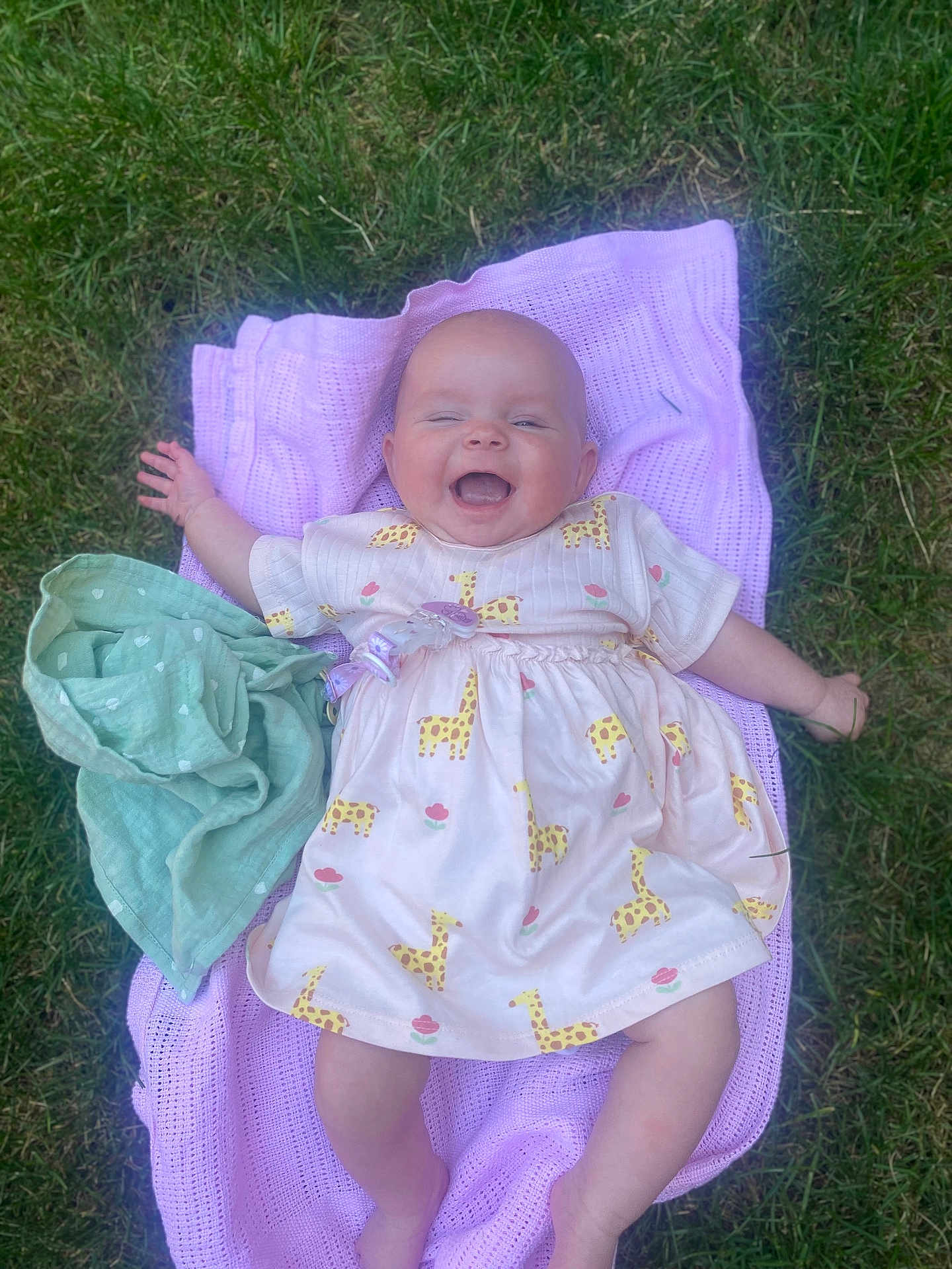 Peach is registered to the contest to win money with this photo: baby, smiling, dress, giraffe_pattern, blanket, grass, outdoor, happy, child, infant, person, cute, lying_down, playful, summer, nature, young, cheerful, clothing, pacifier_clip