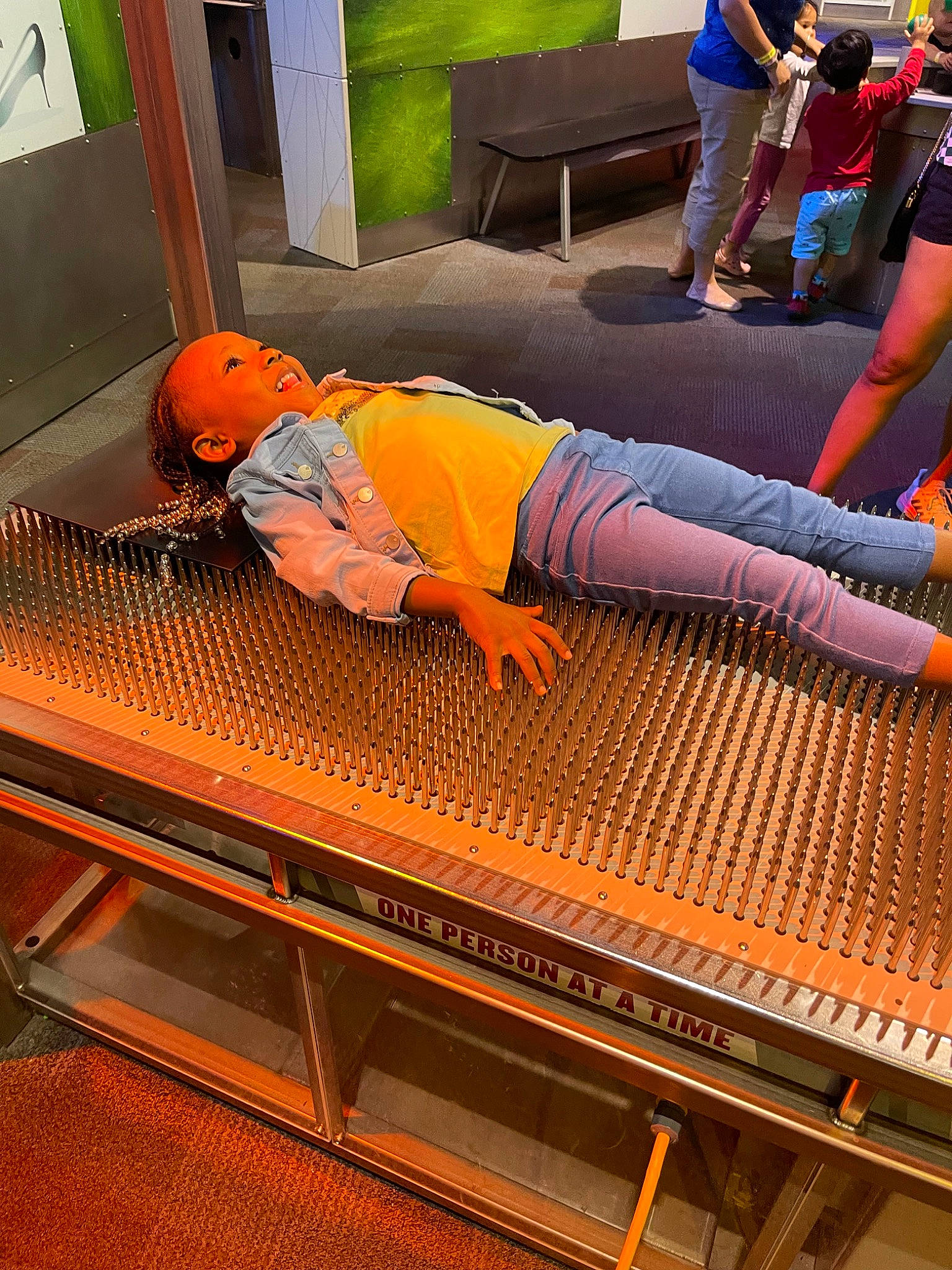 Kinley is registered to the contest to win money with this photo: balance, flooring, fun, human_leg, knee, leisure, machine, metal, orange, pattern, person, physical_fitness, rectangle, room, shorts, t_shirt, thigh, toddler, weaving, wood