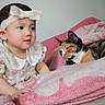 baby, child, headband, dress, pink_blanket, cat, calico_cat, sleeping_cat, indoor, cozy, curious, soft_texture, bed, cuddly, pet, portrait, infant, animal, closeup, cute