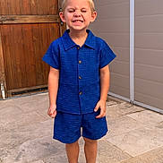 Aïden Kuntz a rejoint le concours — aidez-le/la à gagner de superbes lots ! blue_clothing, boy, casual, child, cute, eyes_closed, happy, outdoor, person, portrait, shirt, shorts, smiling, sneakers, standing, stone_patio, summer, wall, wooden_door, young