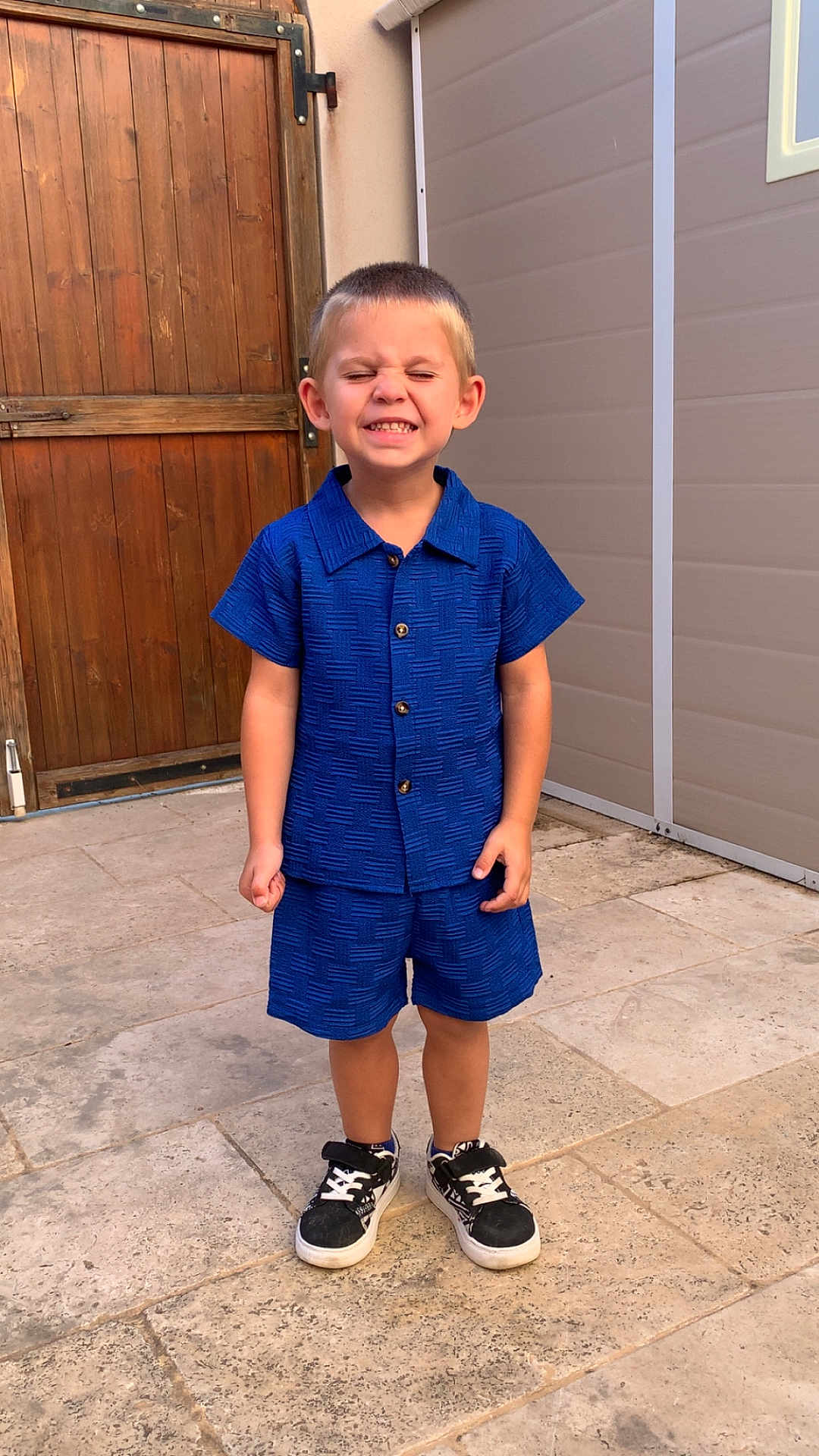 Aïden Kuntz a rejoint le concours — aidez-le/la à gagner de superbes lots ! blue_clothing, boy, casual, child, cute, eyes_closed, happy, outdoor, person, portrait, shirt, shorts, smiling, sneakers, standing, stone_patio, summer, wall, wooden_door, young