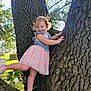 child, toddler, tree, climbing, smiling, pink_tutu, denim_top, shoes, outdoor, nature, sunlight, grass, park, trunk, bark, greenery, happy, playful, young_child, daytime