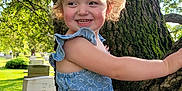 Sarah is registered to the contest to win money with this photo: child, toddler, girl, smiling, tree, nature, outdoor, sunlight, grass, dress, blue_top, pink_skirt, happy, curly_hair, moss, bark, park, greenery, portrait, daylight