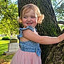 child, toddler, girl, smiling, tree, nature, outdoor, sunlight, grass, dress, blue_top, pink_skirt, happy, curly_hair, moss, bark, park, greenery, portrait, daylight