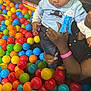 child, toddler, adult, hand, ball_pit, colorful_balls, blue_ribbon, clothing, jeans, nail_polish, play_area, indoor, person, skin, face, holding, pink_wristband, black_shirt, toy, fun