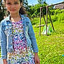 child, girl, outdoor, daylight, grass, dress, butterfly_pattern, denim_jacket, swing_set, greenery, smile, curly_hair, nature, playground, sunny, casual_clothing, portrait, person, happy, blue_sky