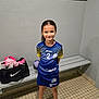 child, girl, sports_uniform, locker_room, bench, sports_bag, handball, smile, pink_shoes, tiled_floor, indoor, portrait, standing, shorts, socks, young, confident, hair_tied_back, team_sport, athlete