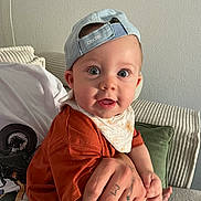 Andrea a rejoint le concours — aidez-le/la à gagner de superbes lots ! baby, child, cap, hat, blue_cap, bib, tattoo, hand, adult_hand, couch, cushion, orange_shirt, yellow_pants, indoor, person, face, wide_eyes, curious, seated, closeup