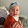 baby, child, cap, hat, blue_cap, bib, tattoo, hand, adult_hand, couch, cushion, orange_shirt, yellow_pants, indoor, person, face, wide_eyes, curious, seated, closeup