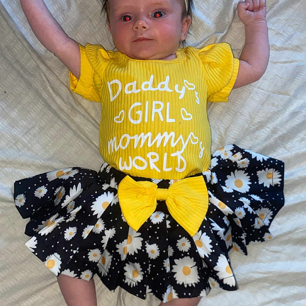 Daisy is registered to the contest to win money with this photo: baby, bed, black_skirt, blanket, child, cute, daisy_pattern, fashion, footwearless, happy, headband, indoors, infant, lying_down, person, portrait, raised_arms, smiling, toddler, yellow_clothing