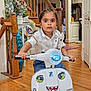 child, toy_scooter, indoor, wooden_floor, staircase, vase, plant, wide_eyes, white_shirt, blue_jeans, mirror, door, hand, floor, window, decor, expression, playful, face_sticker, young