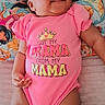 baby, child, infant, smiling, happy, portrait, pink_clothing, onesie, crown_graphic, text_on_clothing, lying_down, bed, pillow, blanket, cartoon, colorful, indoor, nursery, close_up, family_fun