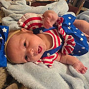 Dakota is registered to the contest to win money with this photo: baby, infant, blue_bow, headband, stars, stripes, red_white_blue, blanket, soft_texture, lying_down, cute, patriotic_outfit, clothing, young_child, person, skin, hand, face, indoors, expression