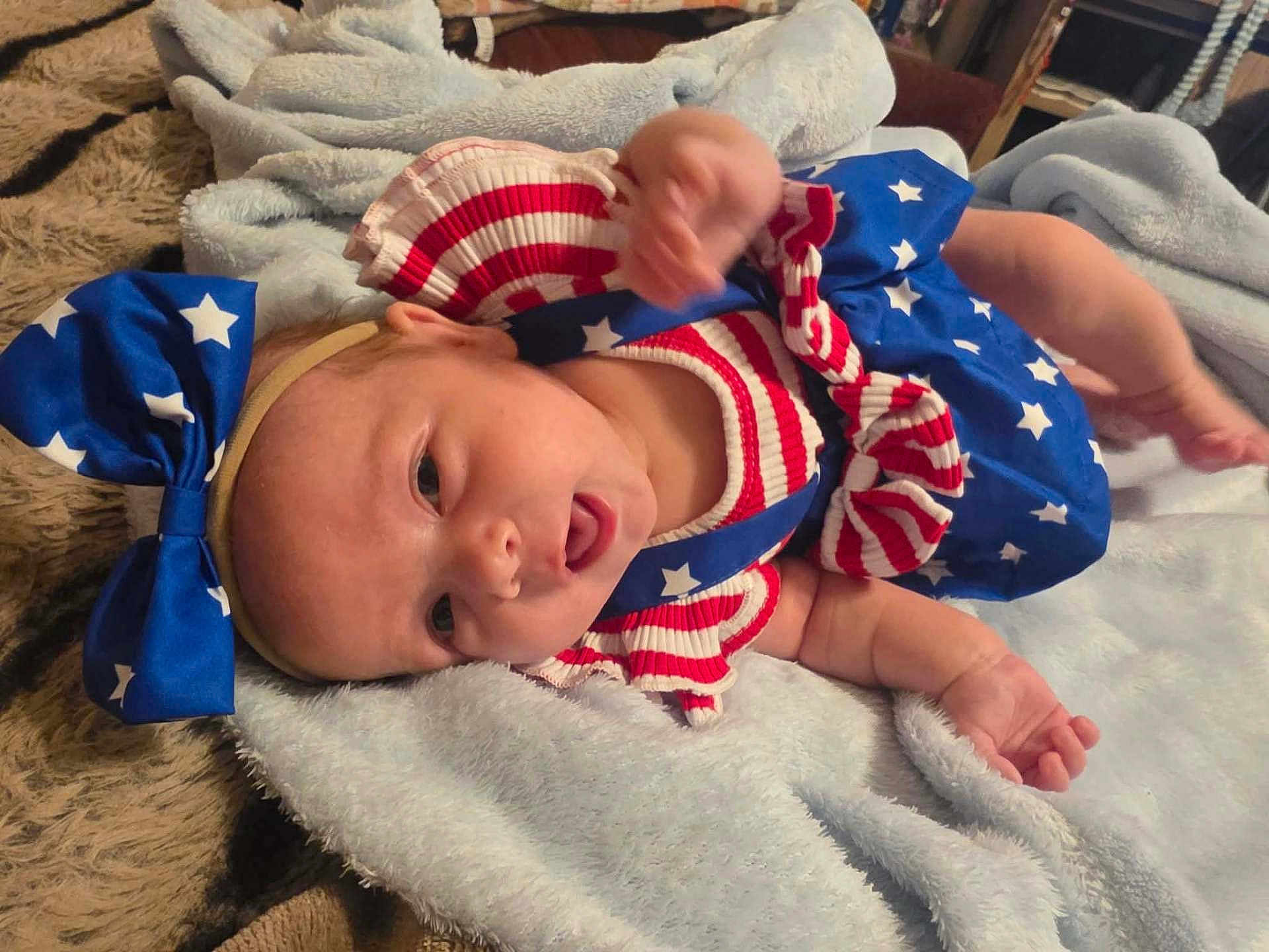 Dakota is registered to the contest to win money with this photo: baby, infant, blue_bow, headband, stars, stripes, red_white_blue, blanket, soft_texture, lying_down, cute, patriotic_outfit, clothing, young_child, person, skin, hand, face, indoors, expression