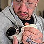 man, glasses, tattoo, hand, kitten, bottle, feeding, black_and_white, hoodie, indoor, closeup, pet, animal, care, cute, young_animal, fur, face, person, holding