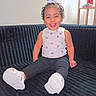Nexielys joined the competition — help win amazing prizes! toddler, child, smiling, sitting, couch, white_shoes, black_pants, white_top, rainbow_pattern, hair_bun, indoor, furniture, shelf, toy, happy, portrait, person, feet, room, home