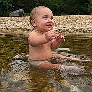 Navy is registered to the contest to win money with this photo: baby, child, water, splashing, smiling, outdoor, nature, rocks, river, trees, happy, bare_chest, summer, playful, cute, sitting, young, natural_light, water_surface, fun